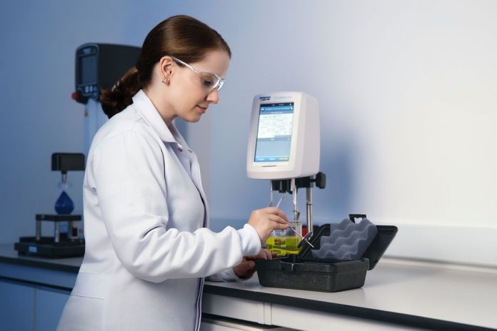 Mulher usando jaleco e óculos de proteção em um laboratório, analisando uma amostra química.