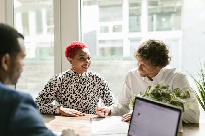 Uma mulher de cabelo curto e vermelho sorri durante uma reunião com dois homens.