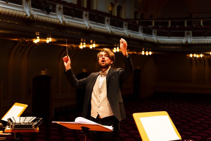 Maestro de fraque conduzindo uma orquestra com a batuta erguida em uma elegante sala de concerto, com estantes de partitura em primeiro plano