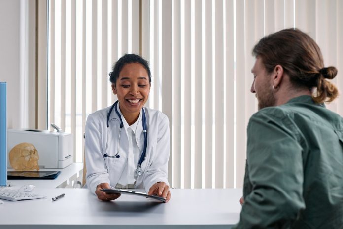Médica de jaleco branco conversa com paciente adulto em consultório claro enquanto registra informações na prancheta